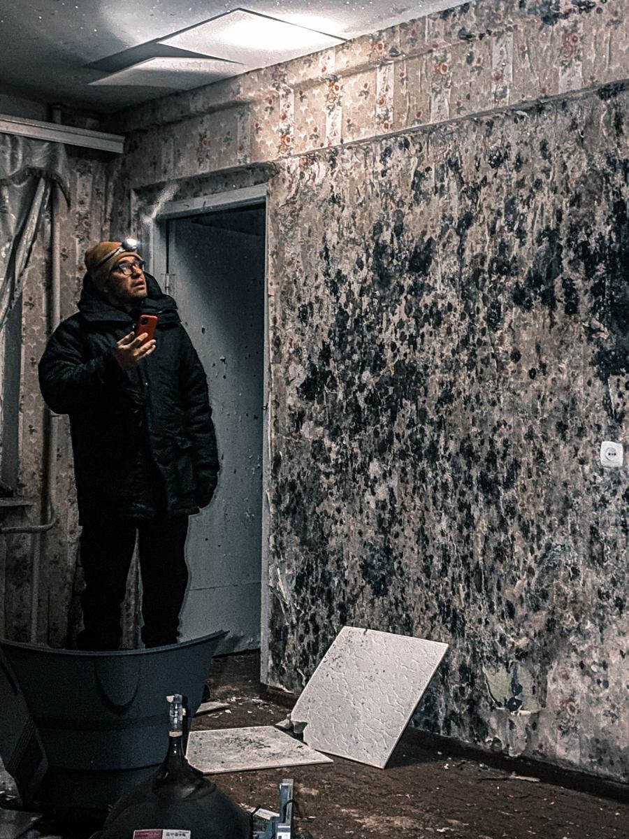 Man with a headlamp inspecting a dark room whose walls are covered in black and white mold growth